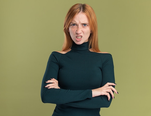 Jeune fille rousse au gingembre en colère avec des taches de rousseur debout avec les bras croisés sur vert olive