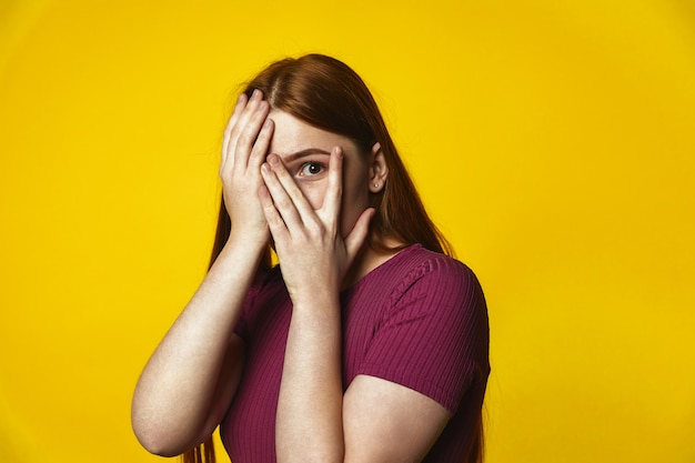 Photo gratuite jeune fille rousse caucasienne couvre le visage avec les mains et regarde à travers les doigts