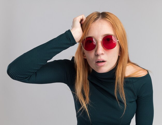 Jeune fille rousse choquée au gingembre avec des taches de rousseur dans des lunettes de soleil met la main sur la tête isolée sur un mur blanc avec espace de copie