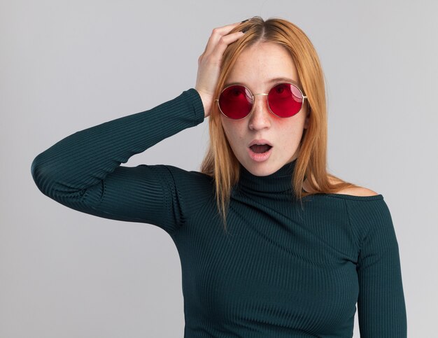 Une jeune fille rousse choquée au gingembre avec des taches de rousseur dans des lunettes de soleil met la main sur la tête et lève les yeux sur un mur blanc avec espace pour copie