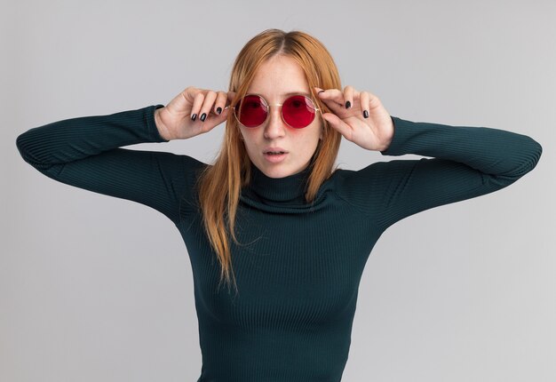 Jeune fille rousse confiante au gingembre avec des taches de rousseur à travers des lunettes de soleil isolées sur un mur blanc avec espace pour copie