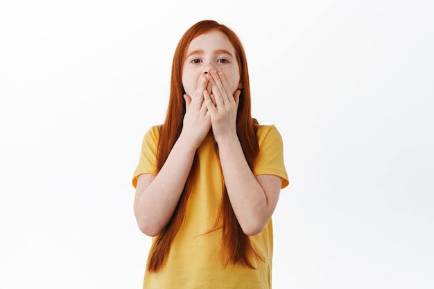 Une jeune fille rousse couvre la bouche et halète, regarde choquée et impressionnée par la caméra, étant étonnée et étonnée, debout en t-shirt jaune sur fond blanc