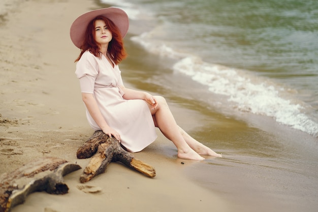 Une jeune fille rousse dans un grand chapeau rond et robe rose assis sur la plage près de l&#39;océan