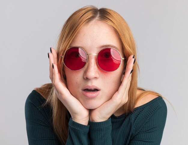 Une jeune fille rousse excitée au gingembre avec des taches de rousseur dans des lunettes de soleil met les mains sur le menton isolé sur un mur blanc avec un espace de copie