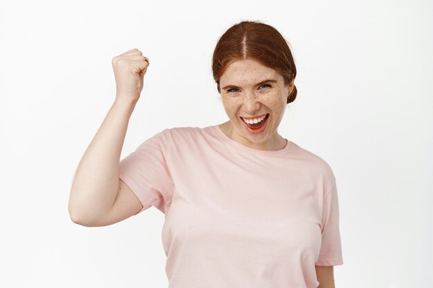 Jeune fille rousse excitée, enracinée pour l'équipe, pompe à poing joyeuse et souriante, déterminée à gagner, acclamant et chantant, gagnant, célébrant la victoire, debout sur blanc.