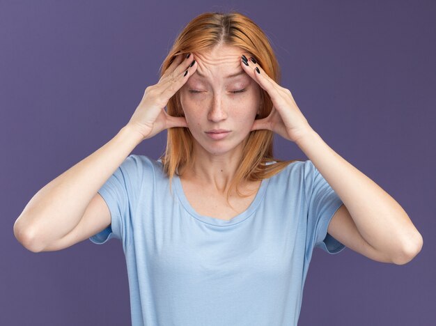 Une jeune fille rousse fatiguée au gingembre avec des taches de rousseur met les mains sur le front isolé sur un mur violet avec espace de copie