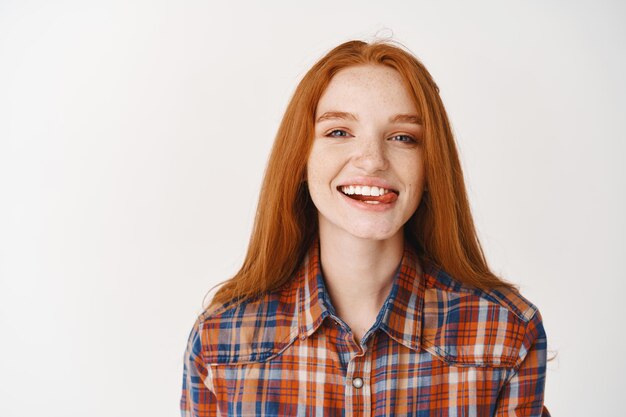 Jeune fille rousse séduisante montrant la langue, souriante avec des dents blanches, l'air insouciant à l'avant, debout sur un mur blanc