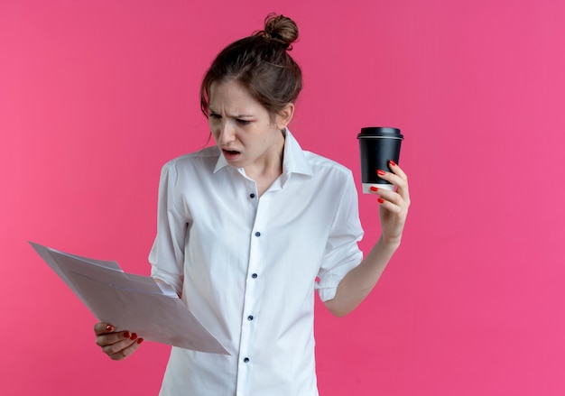 Jeune fille russe blonde agacée regarde des feuilles de papier tenant une tasse de café isolée sur un espace rose avec espace copie