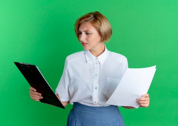 Photo gratuite jeune fille russe blonde confuse détient des feuilles de papier et regarde le presse-papiers isolé sur fond vert avec espace de copie