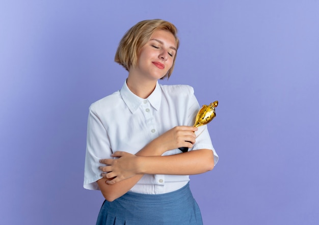 Jeune fille russe blonde heureuse croise les bras et détient la coupe du gagnant isolé sur fond violet avec copie espace