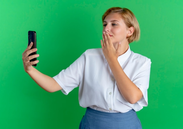 Jeune fille russe blonde heureuse tient et regarde l'envoi de téléphone baiser avec la main isolée sur fond vert avec espace de copie