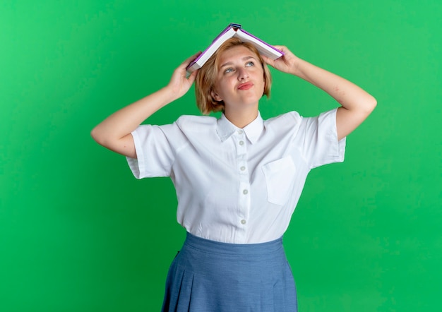 Jeune fille russe blonde joyeuse tient le livre sur la tête isolé sur fond vert avec copie espace