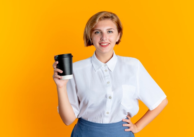 Jeune fille russe blonde souriante tient la tasse de café met la main sur la taille isolé sur fond orange avec copie espace