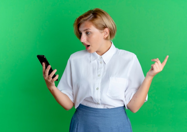 Jeune fille russe blonde surprise regarde le téléphone isolé sur fond vert avec copie espace