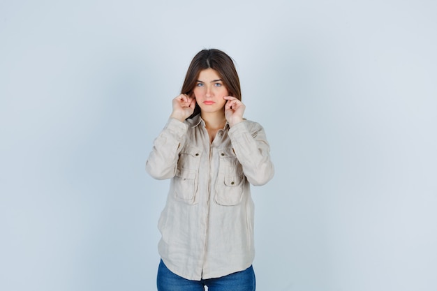 Photo gratuite jeune fille se branchant les oreilles avec l'index en chemise beige, jeans et à l'air contrarié, vue de face.