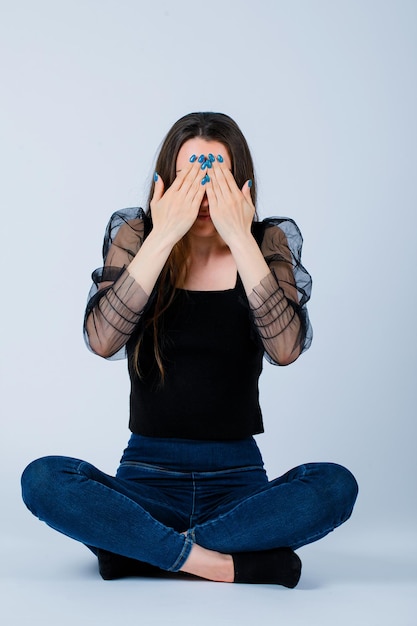 Jeune fille se couvre le visage avec les mains en s'asseyant sur le sol sur fond blanc