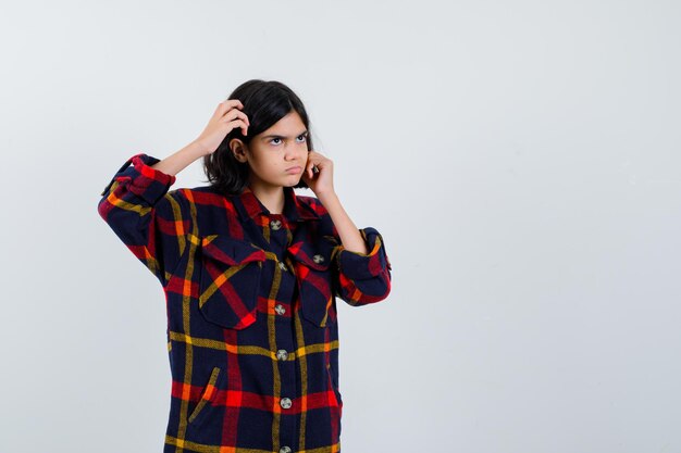 Jeune fille se grattant la tête, mettant la main sur la joue en chemise à carreaux et l'air songeur. vue de face.