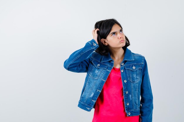 Jeune fille se grattant la tête, pensant à quelque chose en t-shirt rouge et veste en jean et regardant pensive, vue de face.