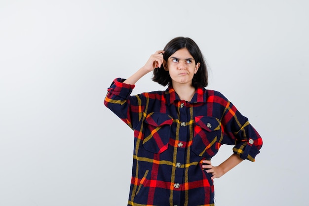 Jeune fille se grattant la tête tout en tenant la main sur la taille en chemise à carreaux et l'air pensif, vue de face.