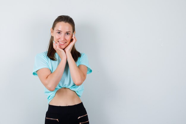 Jeune fille se penchant le menton sur les mains en t-shirt turquoise, pantalon et l'air satisfait, vue de face.