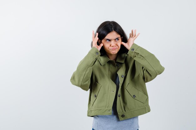 Jeune fille se tenant la main près du visage, regardant loin dans un pull gris, une veste kaki, un pantalon en jean et l'air pensif. vue de face.