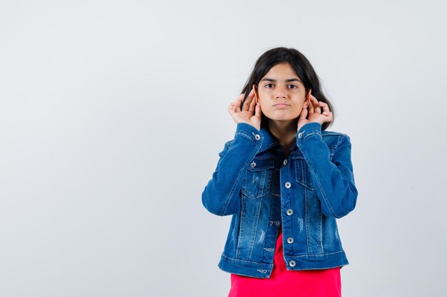 Jeune fille se tenant la main près de l'oreille pour entendre quelque chose en t-shirt rouge et veste en jean et à l'air concentré, vue de face.
