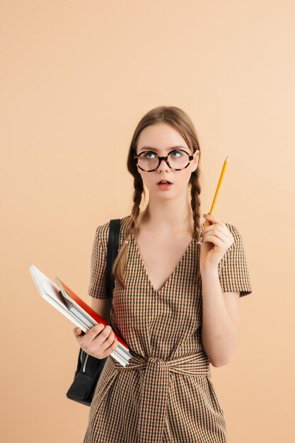 Jeune fille séduisante avec deux tresses en combinaison en tweed et lunettes avec sac à dos noir sur l'épaule tenant un livre à la main et un crayon près de la tête tout en regardant pensivement sur fond beige