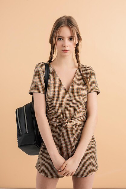 Jeune fille séduisante avec deux tresses en combinaison de tweed avec sac à dos noir sur l'épaule regardant pensivement à huis clos sur fond beige isolé