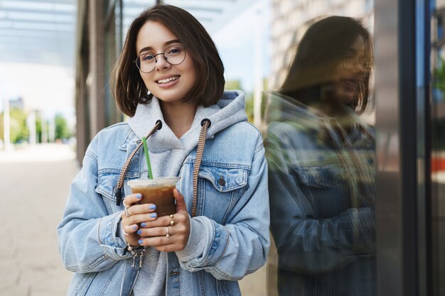 Jeune fille séduisante à lunettes, veste en jean, ayant une promenade décontractée dans la ville, profitant des week-ends, buvant du café au lait, s'appuyant sur le mur du bâtiment et souriant à la caméra avec une expression détendue heureuse.