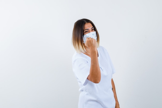 Jeune fille serrant le poing en t-shirt blanc et masque et à la vue de face, confiant.