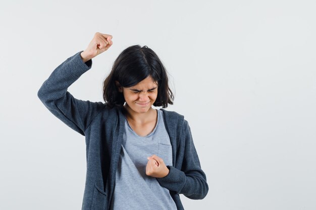 Jeune fille serrant les poings tout en serrant les yeux dans un t-shirt gris clair et un sweat à capuche zippé gris foncé et l'air agacé.