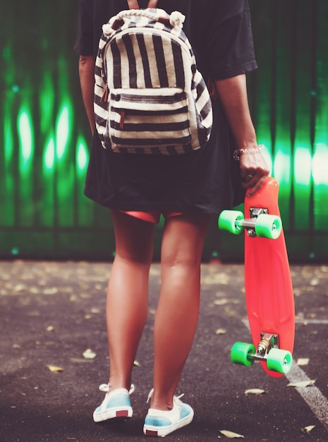 Photo gratuite jeune fille avec shortboard penny orange en plastique derrière le mur vert en casquette
