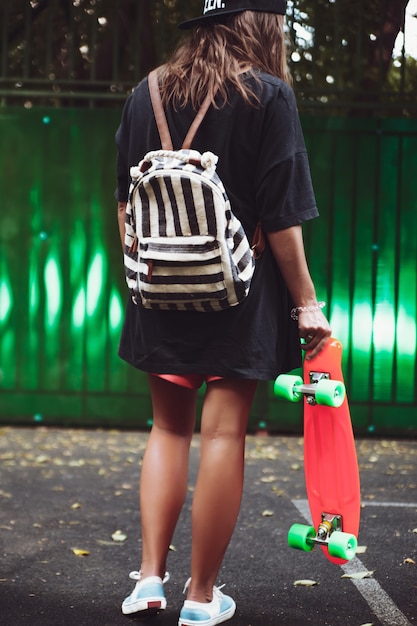 Jeune fille avec shortboard penny orange en plastique derrière le mur vert en casquette