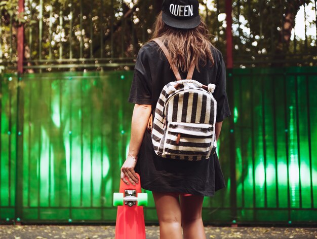 Jeune fille avec shortboard penny orange en plastique derrière le mur vert en casquette