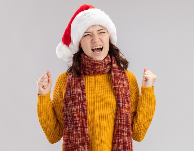 Une jeune fille slave agacée avec un bonnet de noel et une écharpe autour du cou garde les poings et regarde le côté isolé sur un mur blanc avec espace de copie