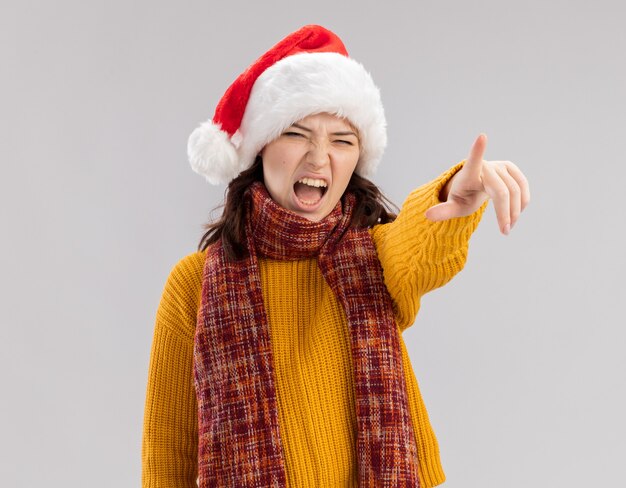 Jeune fille slave agacée avec bonnet de noel et écharpe autour du cou regardant et pointant sur le côté isolé sur mur blanc avec espace de copie