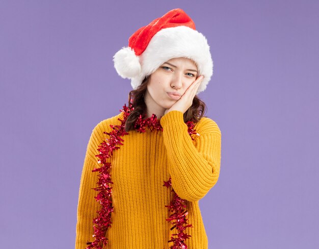 Une jeune fille slave déçue avec un bonnet de noel et une guirlande autour du cou met la main sur le visage isolé sur un mur violet avec espace de copie