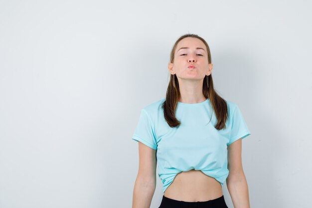 Jeune fille soufflant un baiser d'air en t-shirt turquoise, pantalon et semblant attrayante, vue de face.