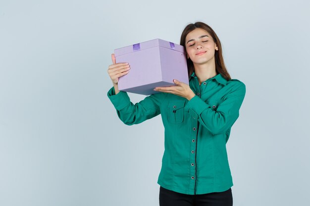 Jeune fille soulevant la boîte-cadeau au-dessus de son épaule en chemisier vert, pantalon noir et à la vue de face, heureux.