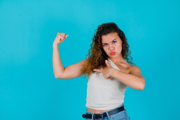 La jeune fille soulève son muscle et le montre avec l'index sur le fond bleu