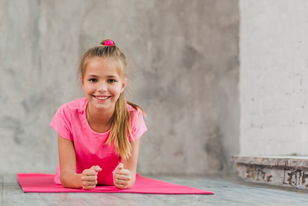 Jeune fille souriante allongée sur un tapis d&#39;exercice rose sur fond de béton