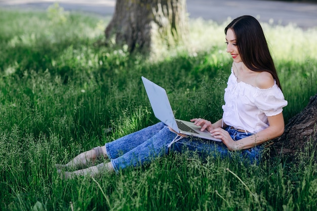 Jeune fille souriante assis sur l&#39;herbe avec un ordinateur portable sur ses jambes