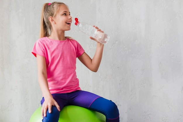 Jeune fille souriante assise sur une balle de pilates verte buvant l’eau de la bouteille contre un mur de béton