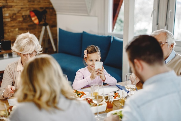 Jeune fille souriante assise à une table à manger avec ses parents et ses grands-parents et utilisant un téléphone portable