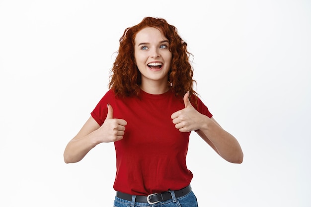 Jeune fille souriante aux cheveux bouclés au gingembre montre les pouces vers le haut et regarde la promotion du produit de louange dans le coin supérieur gauche recommandant la boutique debout sur fond blanc