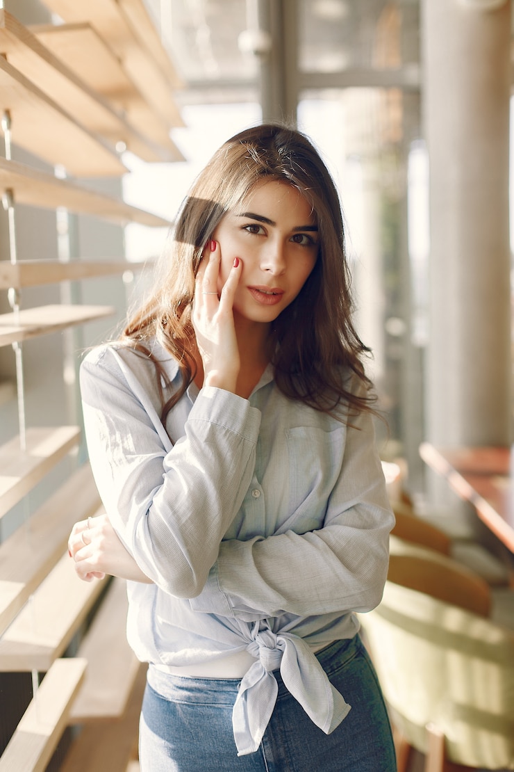 Photo gratuit jeune fille souriante dans une chemise bleue, debout près de la fenêtre et posant
