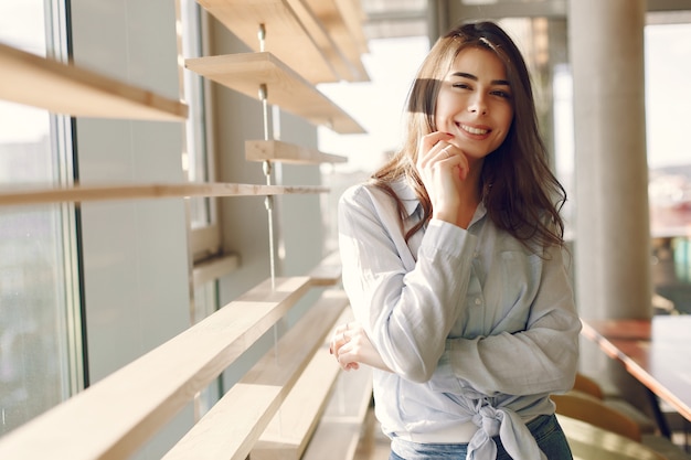 Jeune fille souriante dans une chemise bleue, debout près de la fenêtre et posant