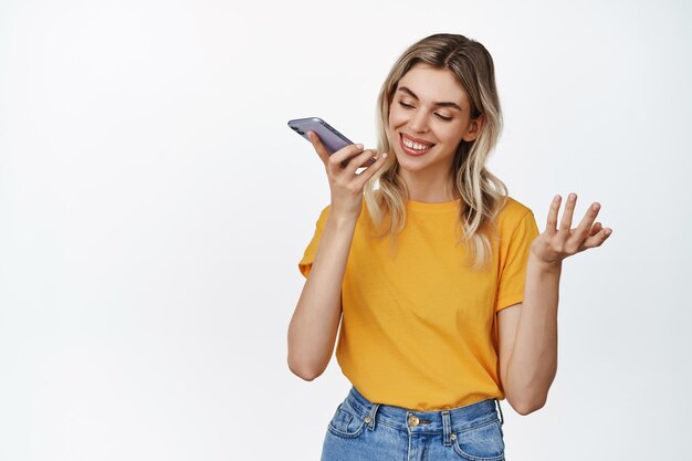 Jeune fille souriante enregistrant sa voix en parlant avec le haut-parleur sur le message vocal d'enregistrement de téléphone portable ou en traduisant ses mots debout sur fond blanc