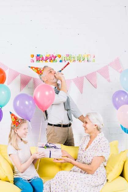 Jeune fille souriante fait un cadeau à sa grand-mère devant l&#39;homme soufflant dans une corne de fête