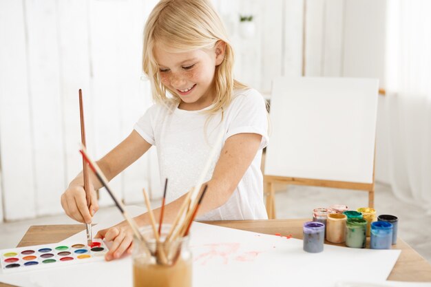 Jeune fille souriante et inspirée avec des cheveux blonds et des taches de rousseur plongeant joyeusement le pinceau dans la peinture rouge, ayant une nouvelle idée pour une photo.